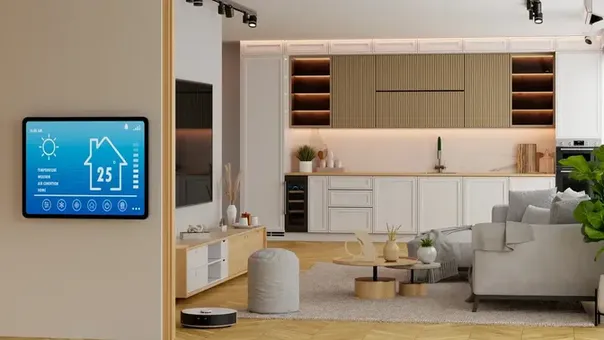 Imagen de gran angular de un salón y cocina modernos, con una pantalla de control de casa inteligente en la pared y un robot aspirador en el suelo.